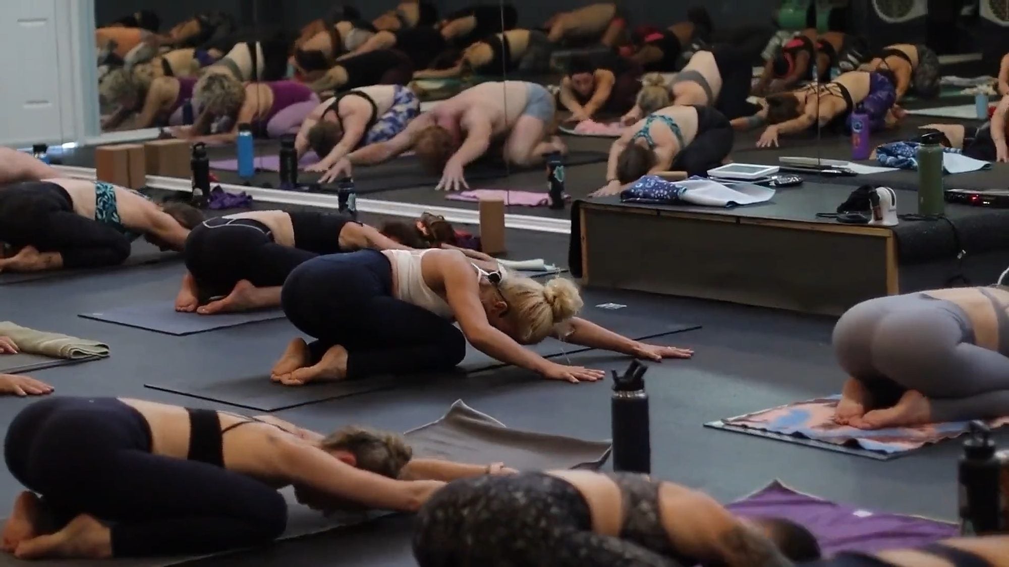 A full hot yoga class folded forward in child pose on dark mats, reflected in the mirror wall